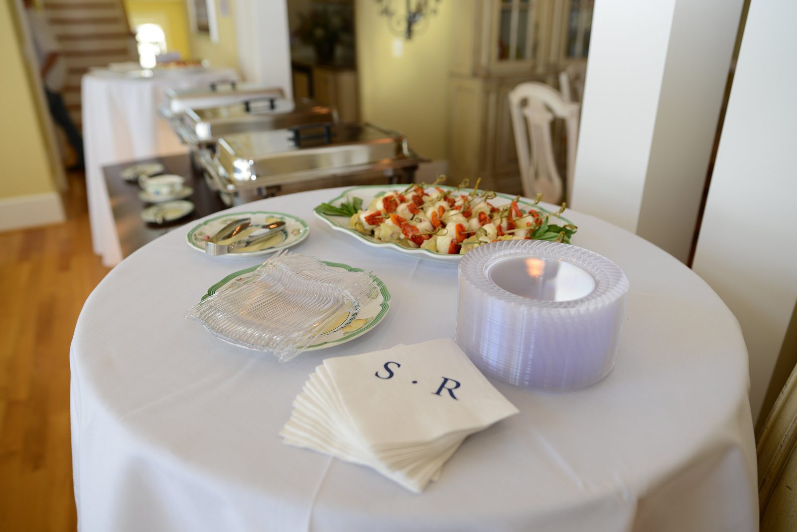 A round table set with linens, plates and a monogrammed napkin stack beside a warming buffet
