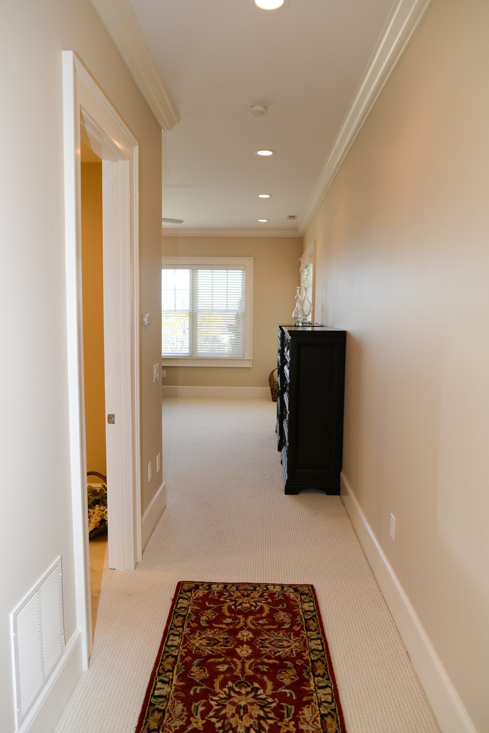 A guest hallway with a runner rug, dresser and natural light at the end