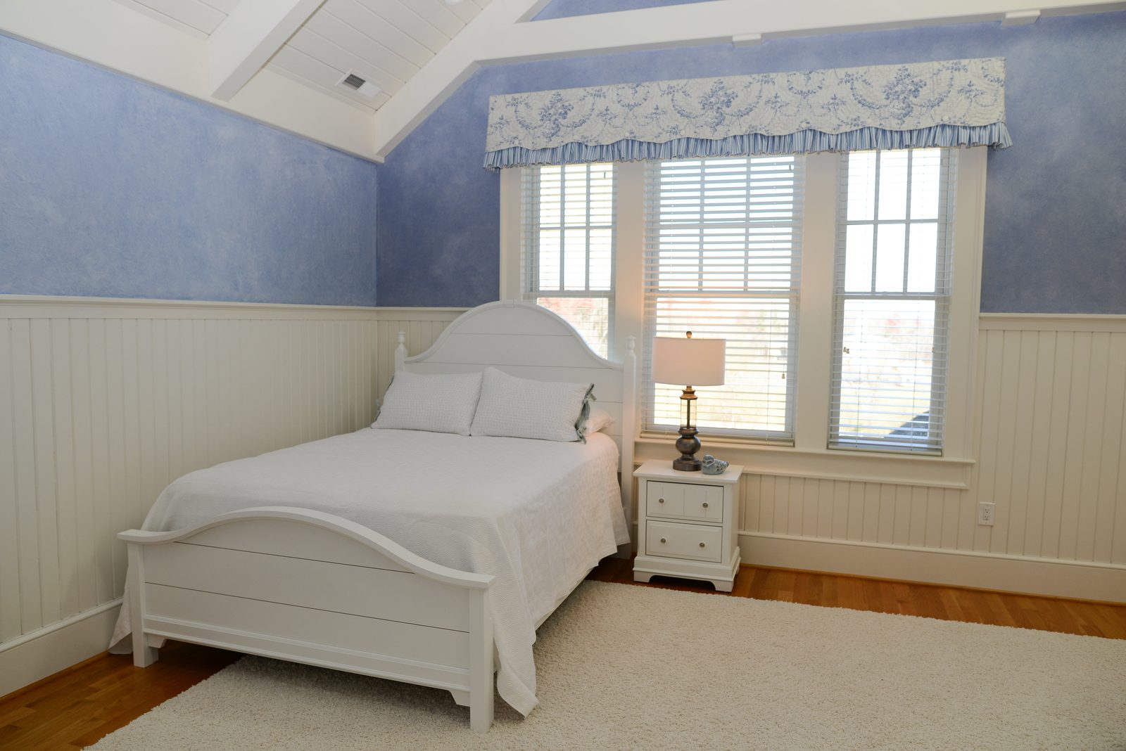 A guest room with a white bed, nightstand and lamp beside a bright window (view 1)