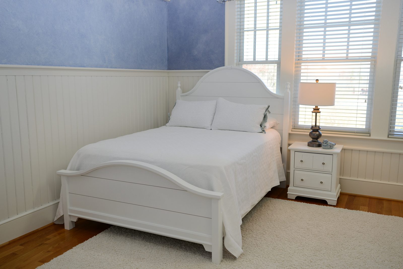 A guest room with a white bed, nightstand and lamp beside a bright window (view 2)