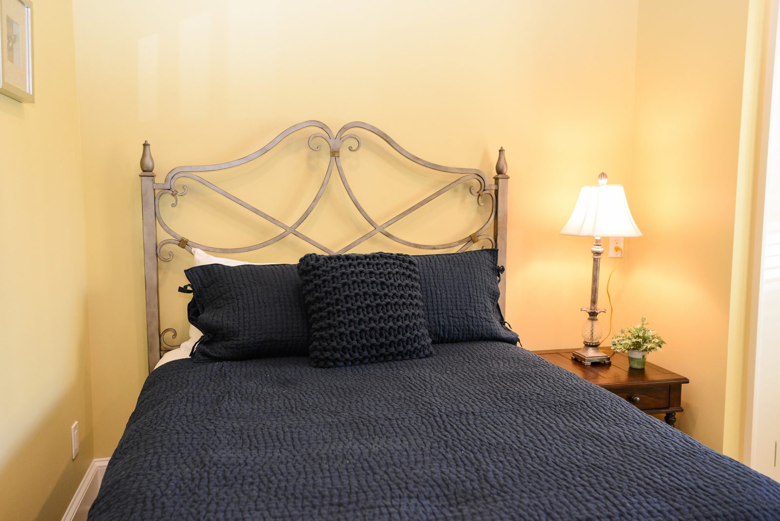 A guest room with an iron bed dressed in navy linens, a lamp and a nightstand (view 1)