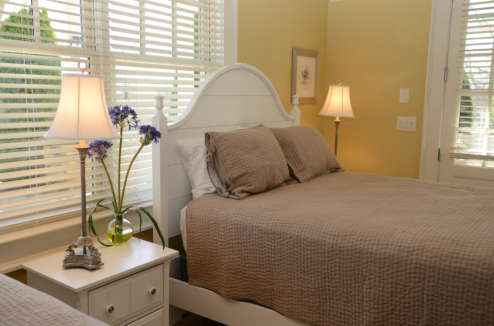 A guest room with a taupe-linened bed, fresh flowers and shuttered windows (view 1)