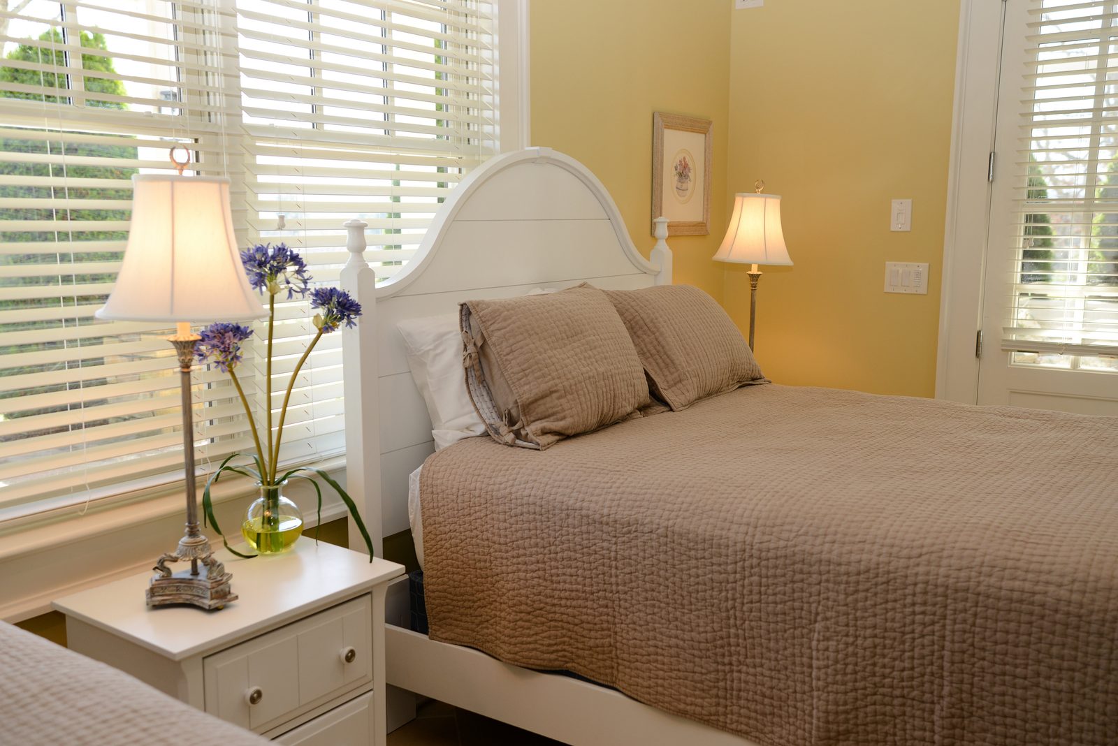 A guest room with a taupe-linened bed, fresh flowers and shuttered windows (view 2)
