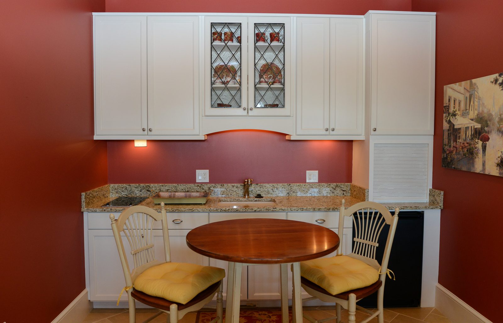 A kitchenette with white cabinetry, a red accent wall and a round cafe table