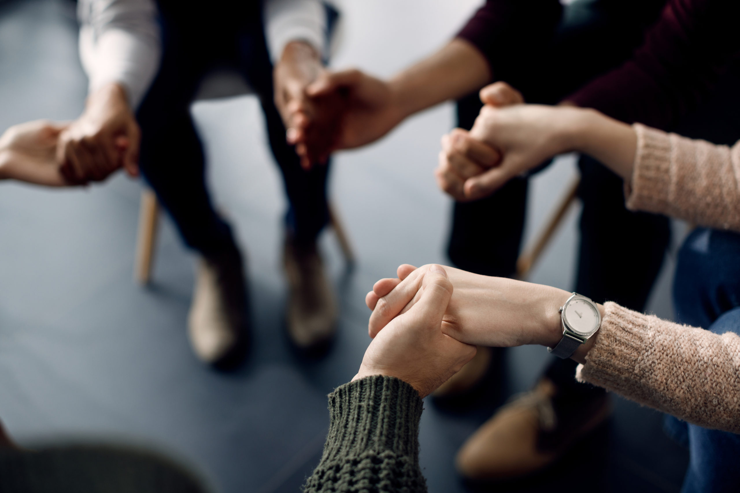 Close-up of people holding hands while sitting in a circle during group therapy.