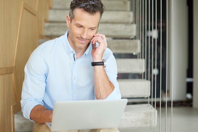Adult male sitting on the steps taking a work call with his laptop on his lap