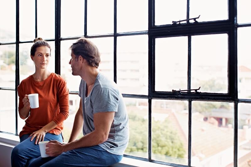 An adult male and female sitting on window ledge with mugs of coffee