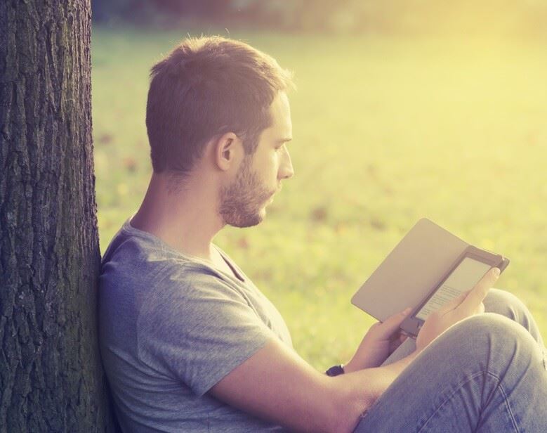 Books-About-Addiction-Recovery Young adult male sitting in field leaned up against a tree reading on his kindle