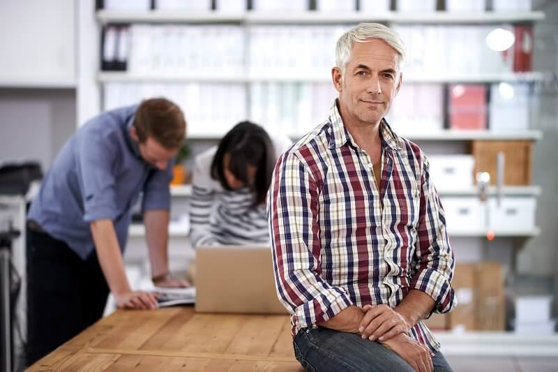 Older adult male sitting on the end of a table in the workplace
