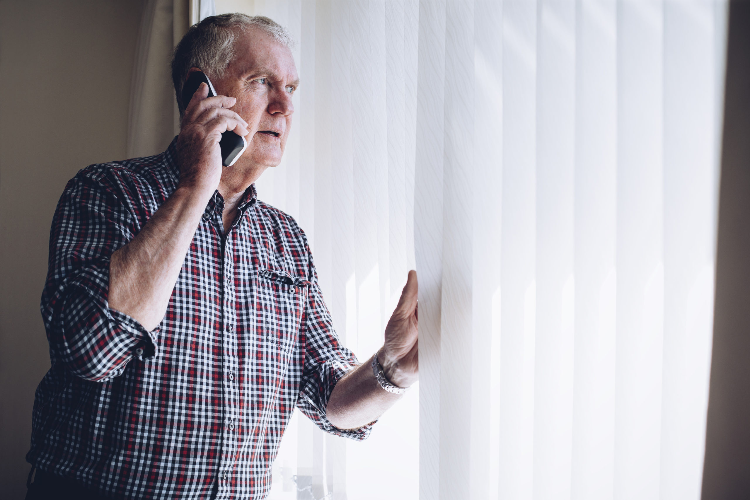 Senior man talking on the phone whilst looking out of his window. He has a worried look on his face.