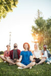 Time for yoga, Attractive young multicultural friends meditating in yoga lotus pose while resting after a long trip in Botanical garden. Multiethnic people dressed in casual summer outdoor clothing.