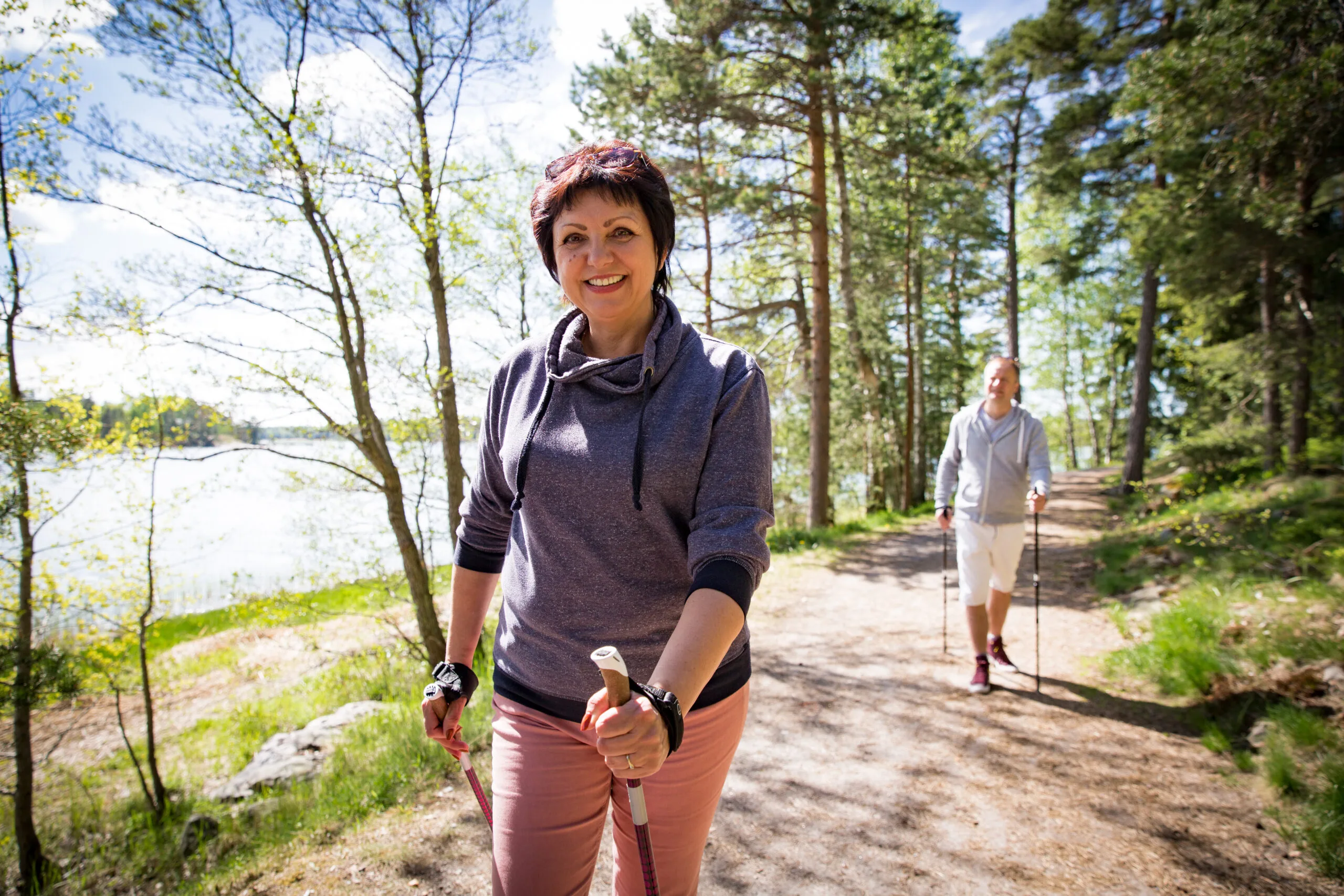 Summer sport in Finland - nordic walking. Man and mature woman hiking in green sunny forest. Active people outdoors. Scenic peaceful Finnish summer landscape.