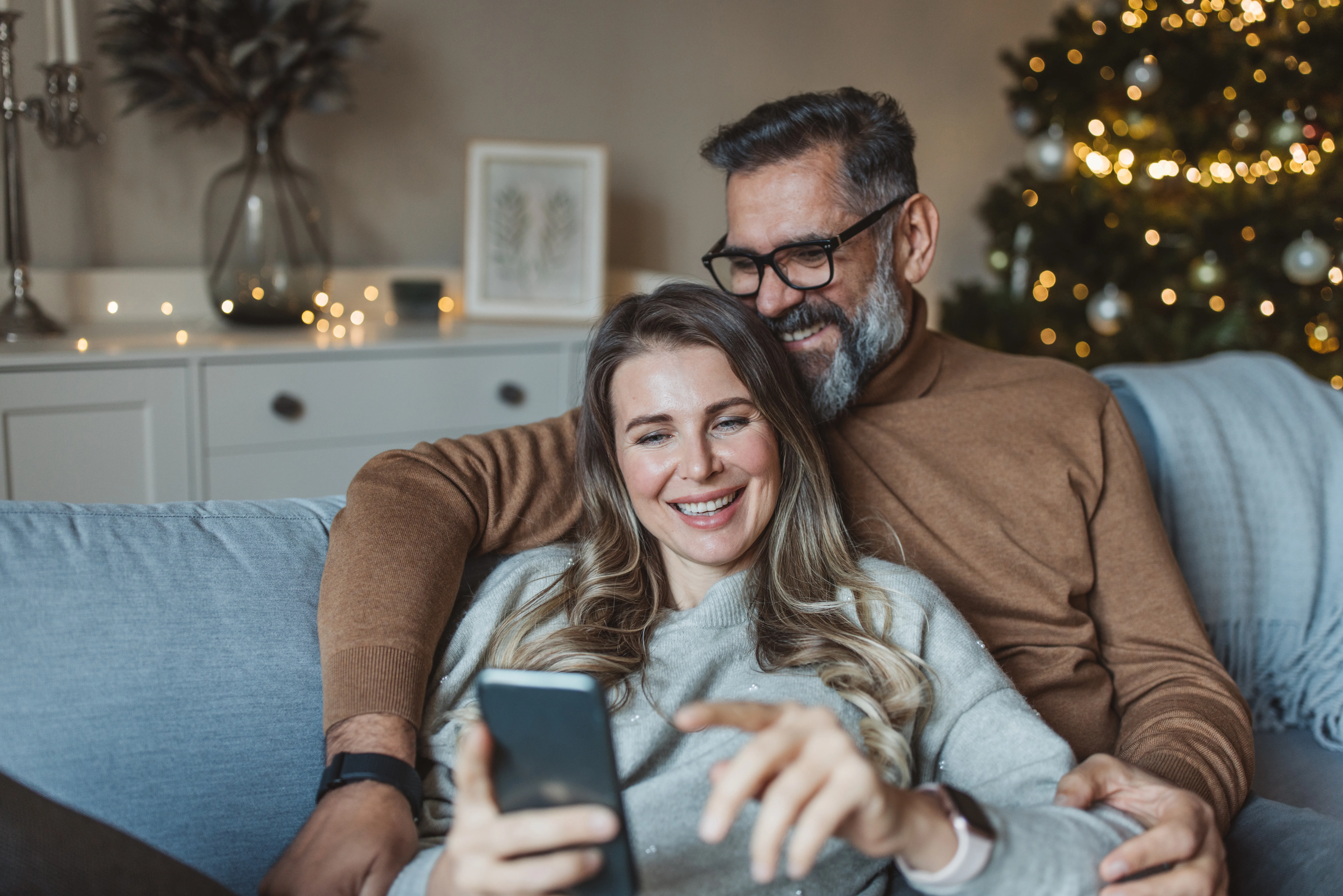 Mature couple sitting on sofa at home and enjoying in there time together. They are shopping online while drinking coffee or tea. It is Christmas time.