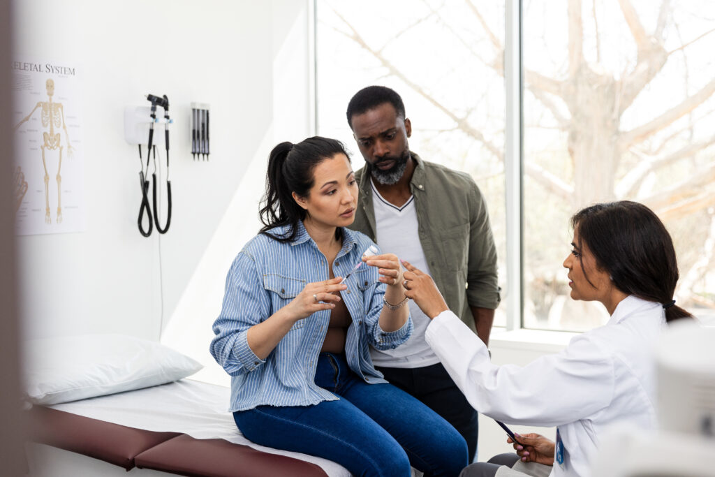 The mid adult female doctor points as she corrects the mid adult woman filling the syringe. The mid adult husband is watching quietly.