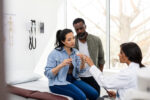 The mid adult female doctor points as she corrects the mid adult woman filling the syringe. The mid adult husband is watching quietly.