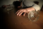Man laying face down on table with hands spread, glass and bottle of alcohol knocked over