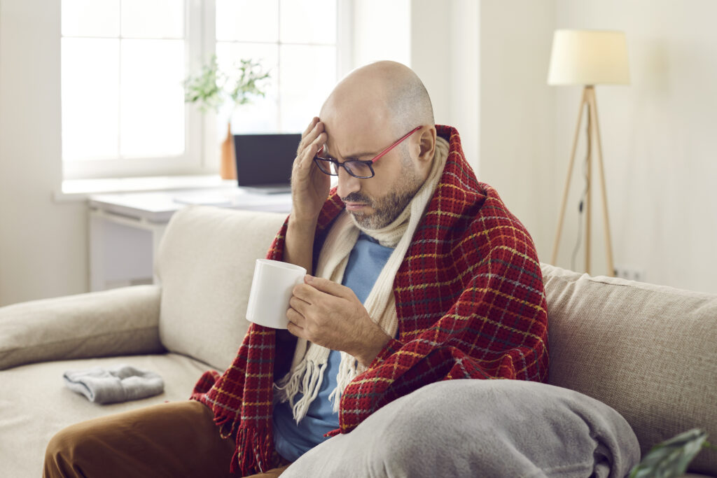 Unhappy joyless sick man at home treats flu and colds with medicinal drink with unpleasant taste. Middle-aged man wrapped in warm scarf and plaid looks at cup in his hands, not wanting to drink it.