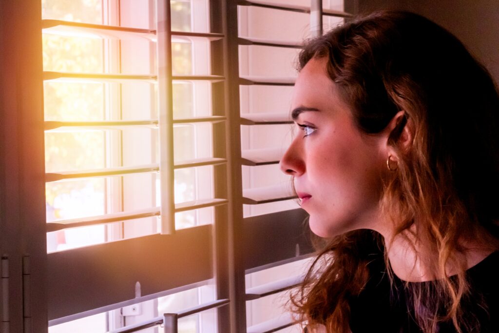 Serious Young woman looking outside through her home window