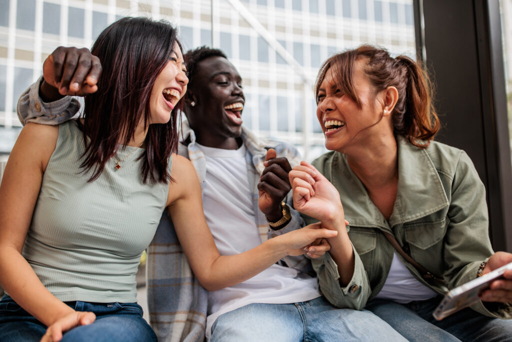 Group of friends from different cultural backgrounds sits together at a bus stop in downtown Sydney, engaging with their smart phone and enjoying a fun, casual moment in the heart of the city.